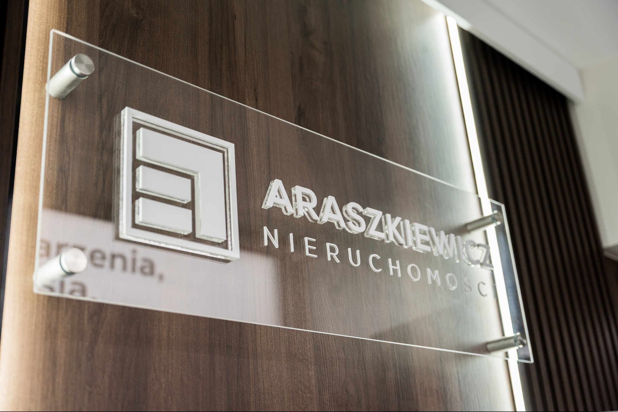 Tabliczka z logo i nazwą 'Araszkiewicz Nieruchomości' na drewnianej ścianie z oświetleniem, zamocowana na metalowych dystansach. Elegancki design.