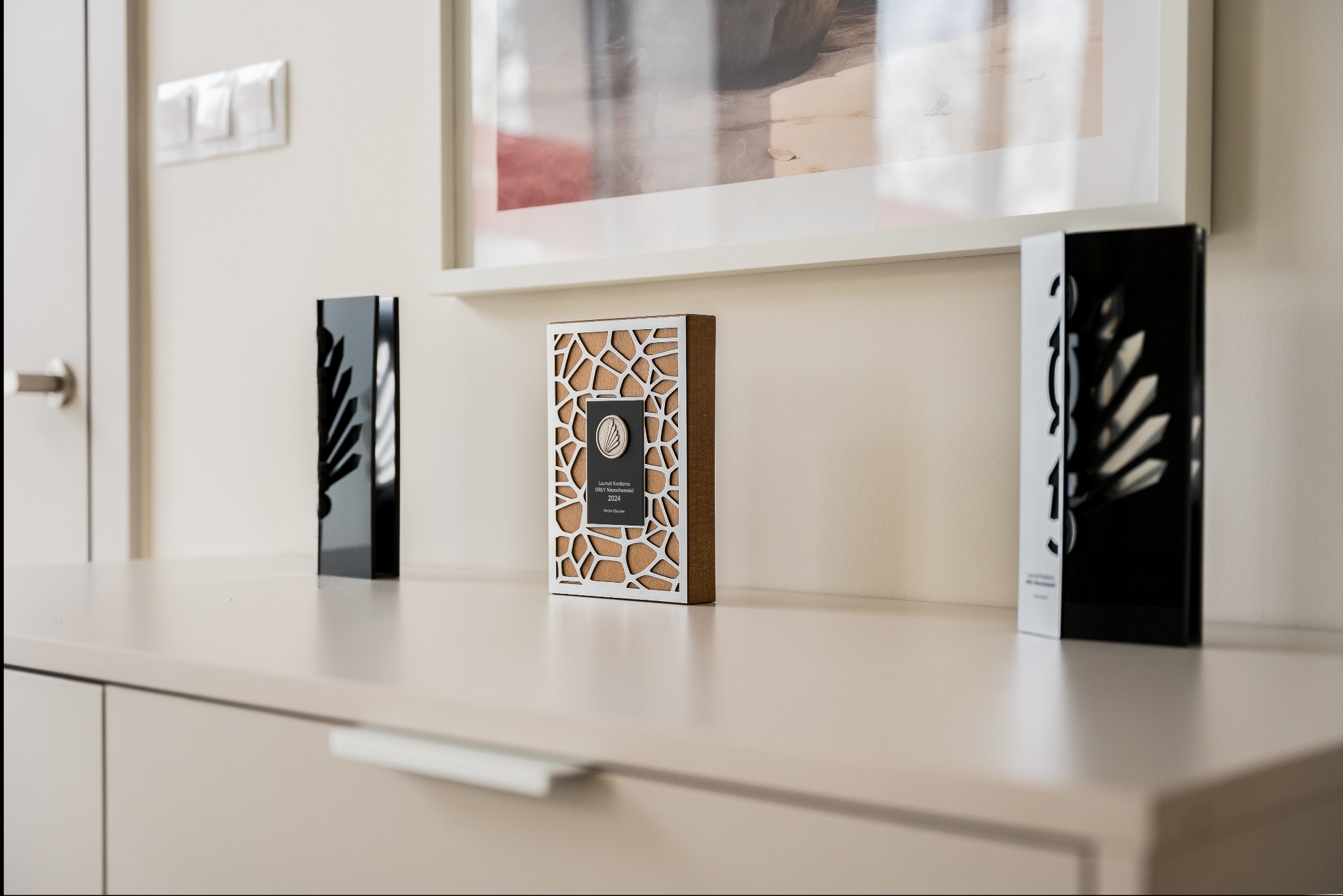 Nagrody 'Orły Nieruchomości 2024' na komodzie w Poznaniu. Statuetki w kolorach czerni, bieli i drewna, z motywem liści, ustawione obok siebie. W tle obraz w białej ramie.