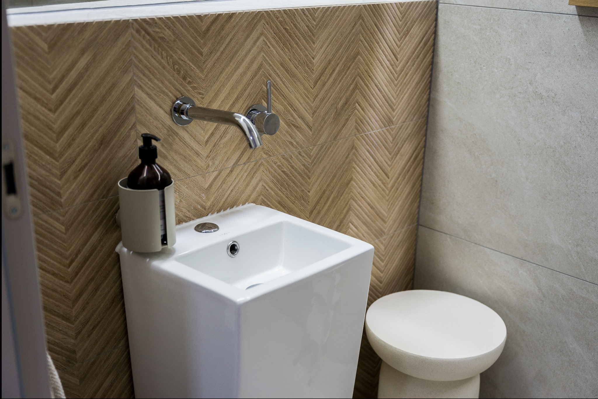 Nowoczesna, biała umywalka z armaturą i dozownikiem mydła na tle ściany z drewnopodobnych płytek ułożonych w jodełkę. Minimalistyczny stołek obok.