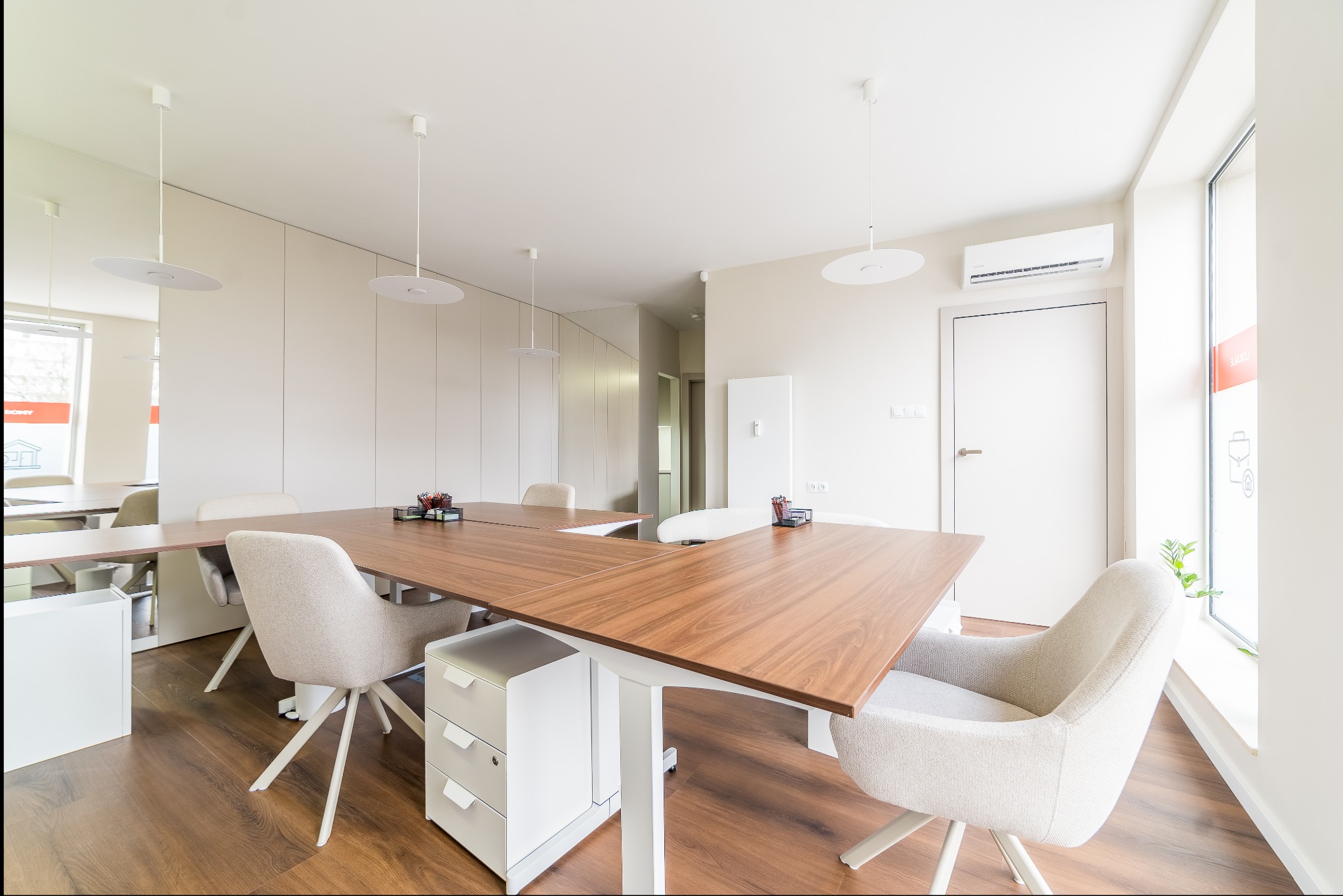 Jasne biuro z drewnianym stołem, krzesłami na kółkach i białymi szafkami. Minimalistyczny design z okrągłymi lampami i drewnianą podłogą. Widok na okno z naklejką.