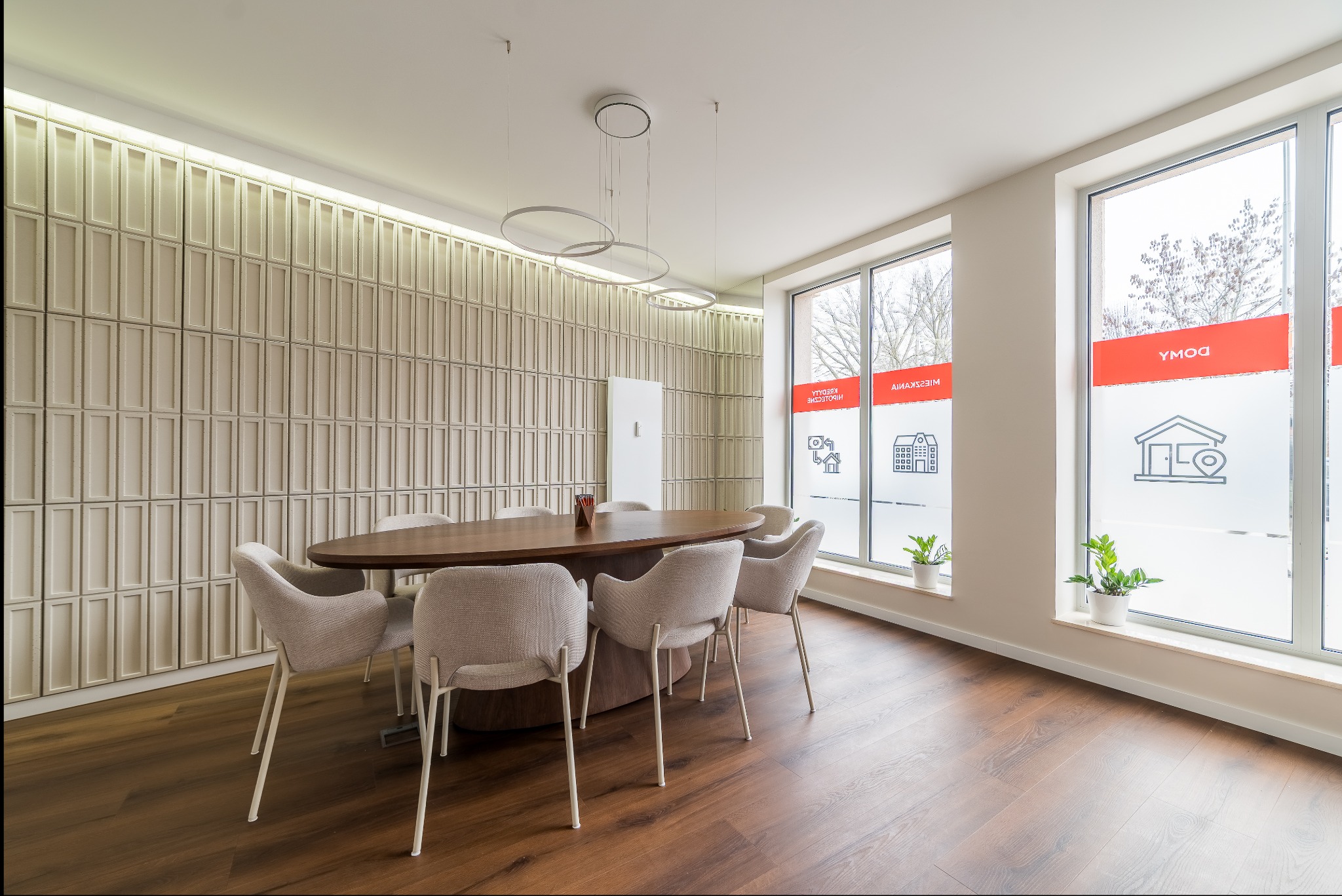 Sala konferencyjna agencji nieruchomości z owalnym stołem, krzesłami i oknami z naklejkami informacyjnymi. Parkiet na podłodze, dekoracyjna ściana.