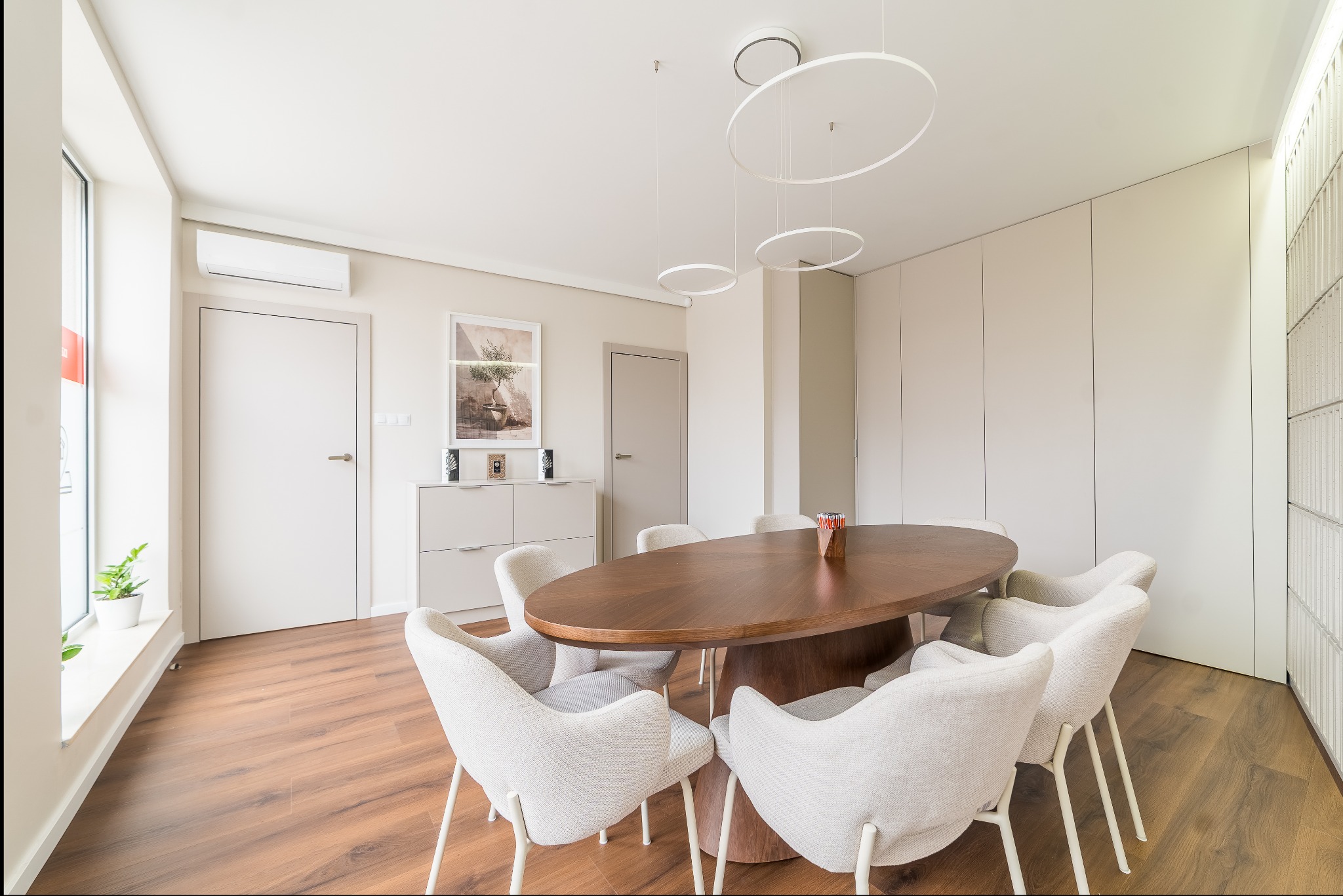 Jasne wnętrze jadalni z owalnym, drewnianym stołem otoczonym krzesłami w odcieniach beżu. Minimalistyczne oświetlenie i drewniana podłoga dodają elegancji.