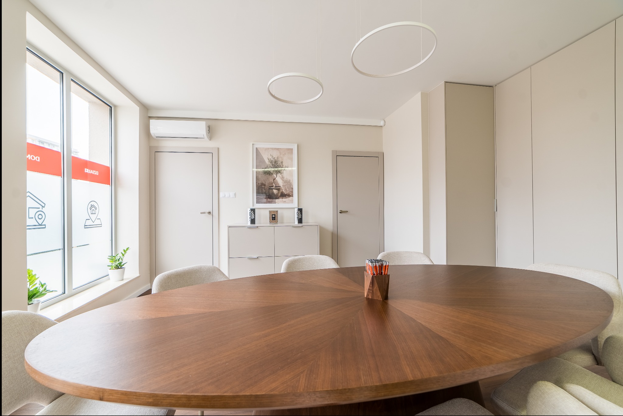 Jasne biuro agencji nieruchomości z owalnym stołem, krzesłami i oknem z logo firmy. Minimalistyczny wystrój z okrągłymi lampami i roślinami w doniczkach.