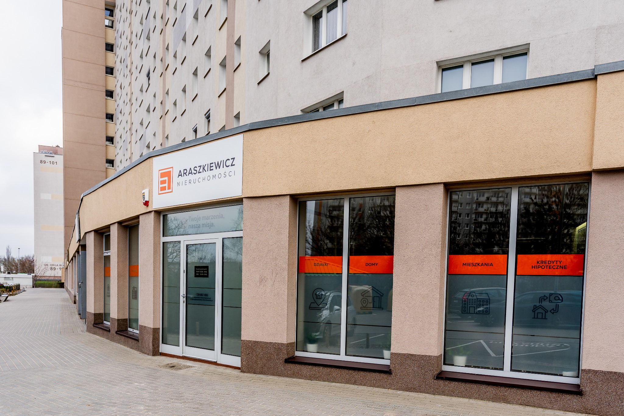 Fasada biura nieruchomości 'Araszkiewicz Nieruchomości' z witrynami oklejonymi grafikami ofert: działki, domy, mieszkania i kredyty hipoteczne. Budynek mieszkalny w tle.