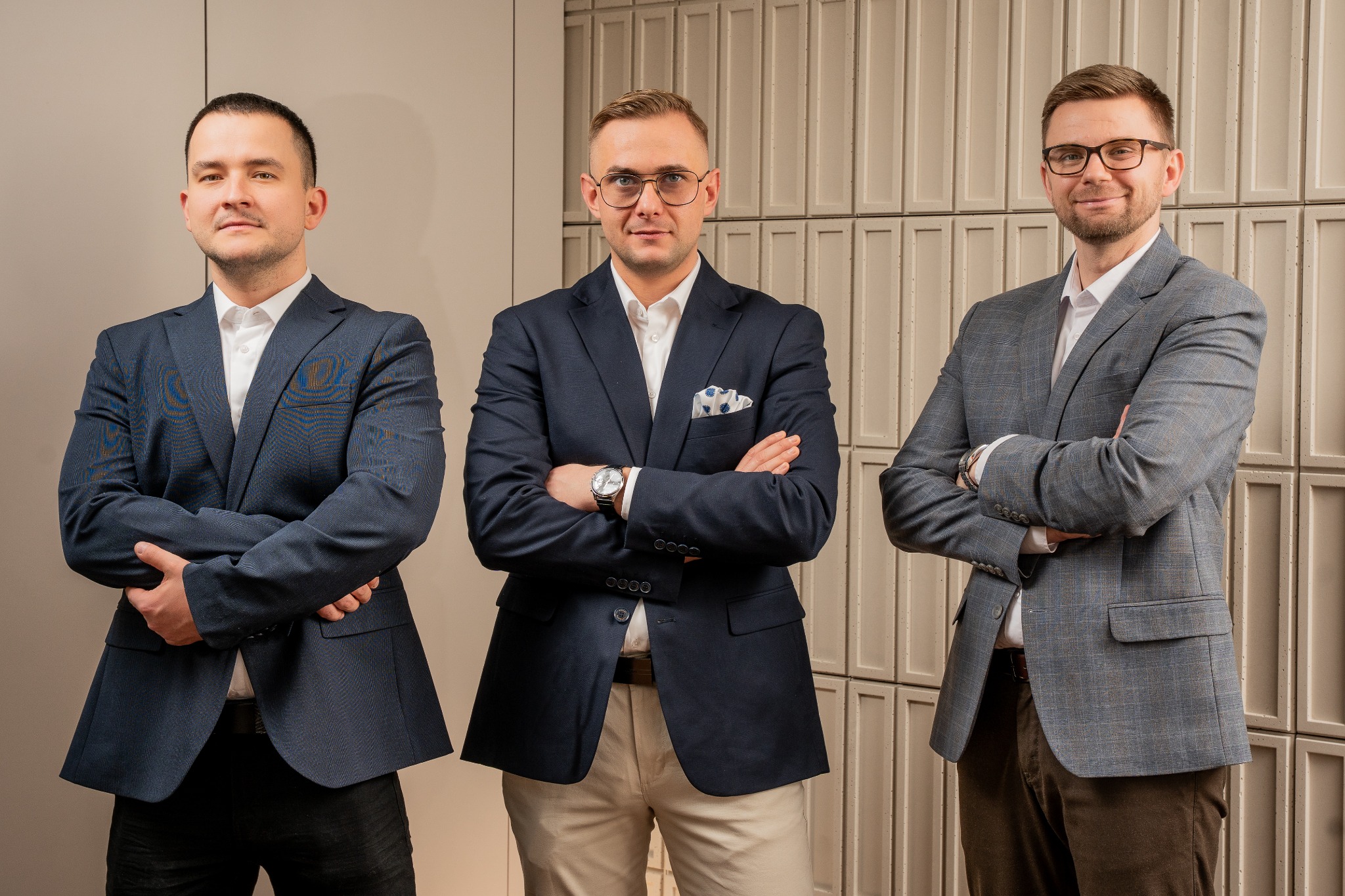 Trzech eleganckich mężczyzn w marynarkach, z założonymi rękami, prezentuje zespół agencji nieruchomości na tle ściany z dekoracyjnymi panelami.