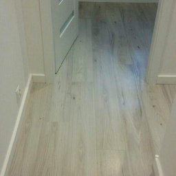 Rafał Smaza - Jasne, drewnopodobne panele podłogowe w korytarzu, widoczne białe listwy przypodłogowe, fragment otwartej białej drzwi z trzema poziomymi przeszkleniami i fragment okna z dekoracją.