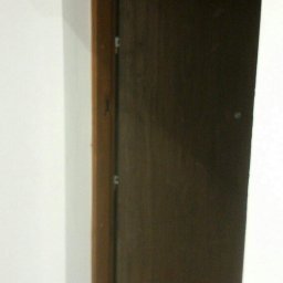 Rafał Smaza - Ciemnobrązowe drzwi z drewnianą ościeżnicą w białej ścianie, widoczne zawiasy.