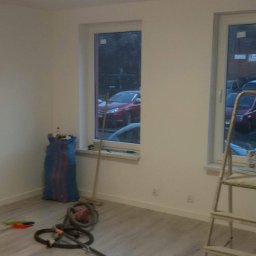 Rafał Smaza - Wnętrze pokoju po malowaniu ścian na biało, widoczne dwa okna z widokiem na parking, drabina, mop, worek na śmieci i narzędzia malarskie na podłodze.