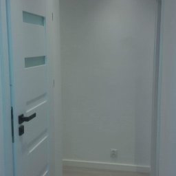 Rafał Smaza - Nowoczesne wnętrze przedpokoju z białymi ścianami, panelami podłogowymi imitującymi drewno i białą, częściowo przeszkloną ościeżnicą drzwiową z czarną klamką