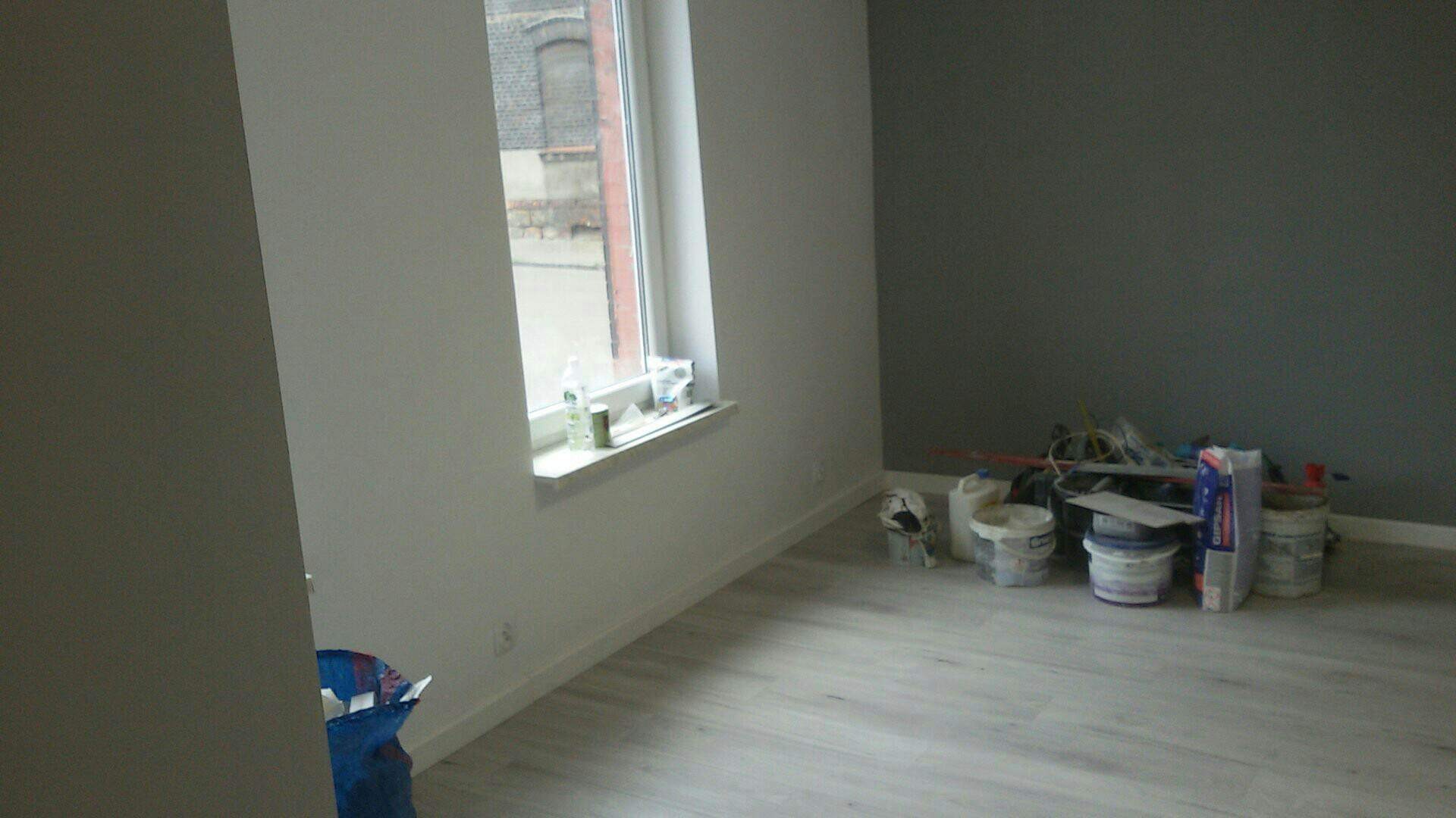 Świeżo pomalowane na biało i szaro ściany w pokoju, z widocznymi narzędziami i materiałami malarskimi przy ścianie, pod oknem, na podłodze z paneli.