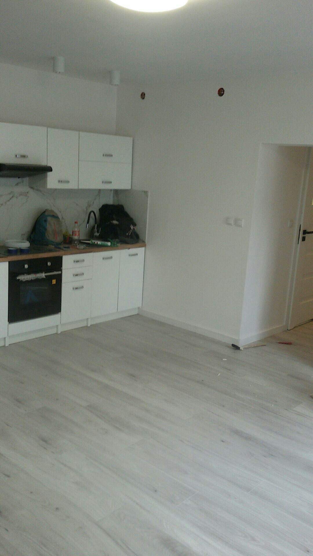 Wnętrze kuchni w trakcie wykańczania: białe szafki kuchenne z marmurowym panelem, jasna podłoga imitująca drewno, surowe ściany z widocznymi otworami na instalacje elektryczne, minimalizm i prostota.