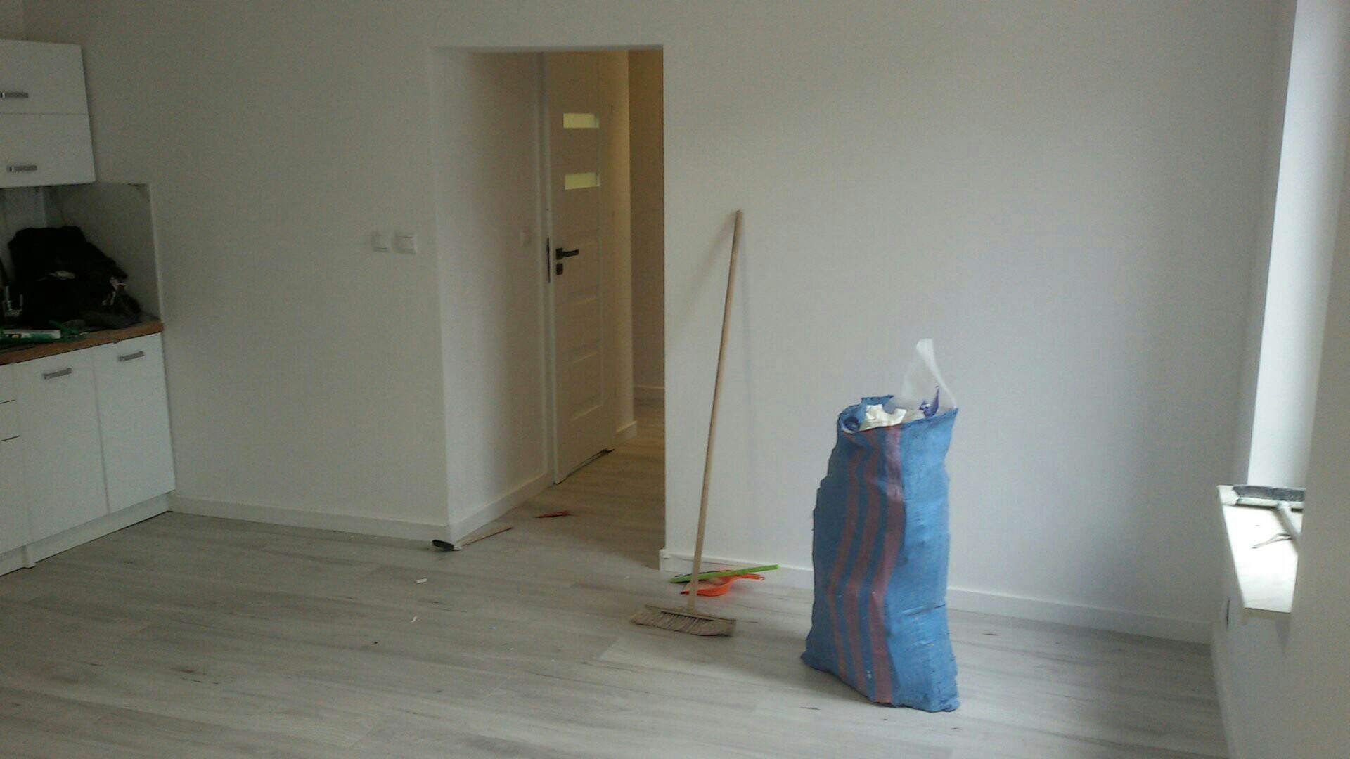 Pokój po remoncie: świeżo pomalowane ściany w odcieniu bieli, jasna podłoga imitująca drewno, widoczny fragment zabudowy kuchennej, otwarte drzwi do kolejnego pomieszczenia oraz worek z odpadami...