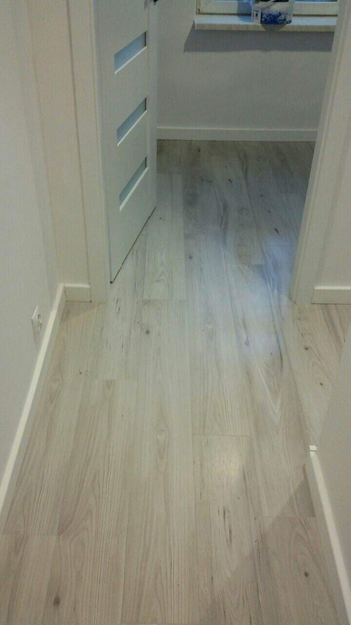 Jasne, drewnopodobne panele podłogowe w korytarzu, widoczne białe listwy przypodłogowe, fragment otwartej białej drzwi z trzema poziomymi przeszkleniami i fragment okna z dekoracją.