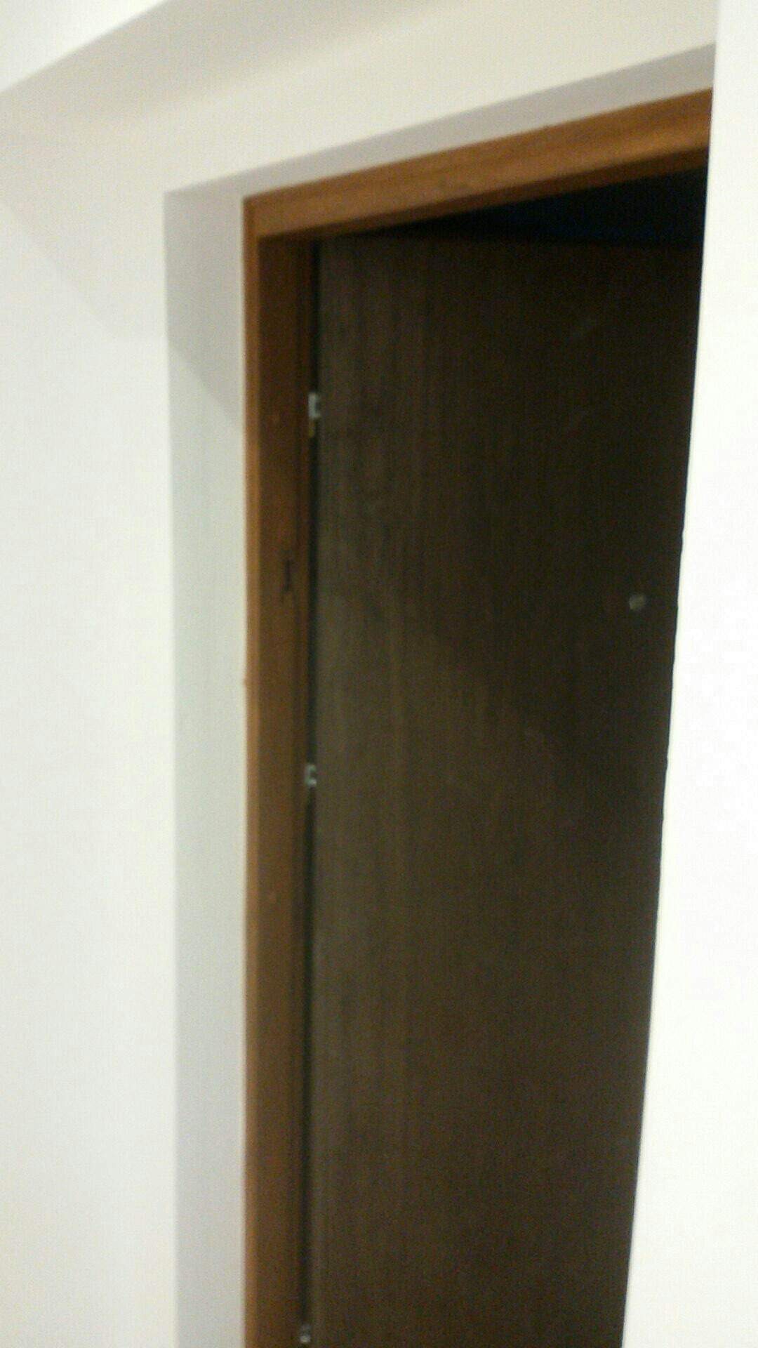 Ciemnobrązowe drzwi z drewnianą ościeżnicą w białej ścianie, widoczne zawiasy.