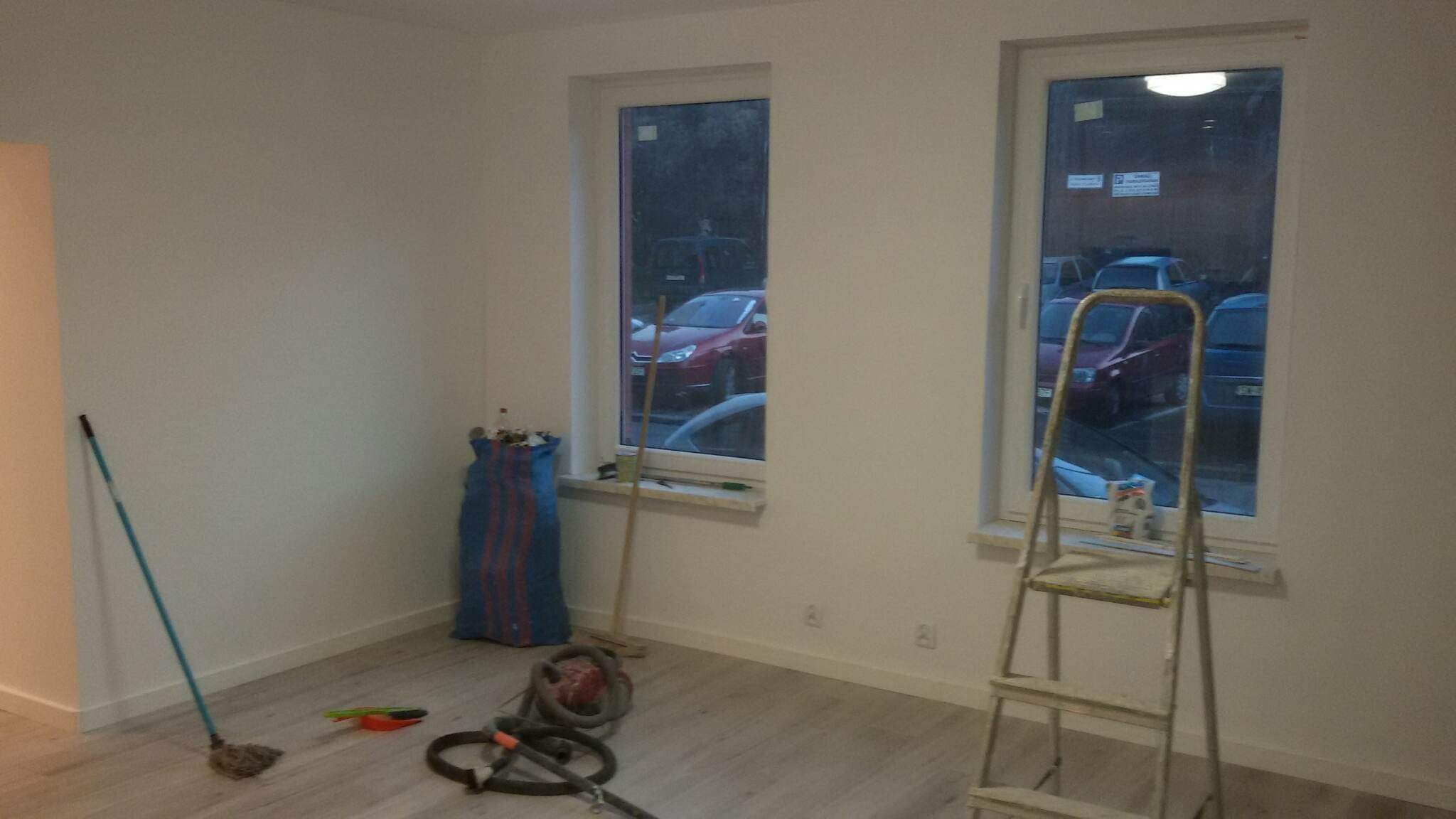Wnętrze pokoju po malowaniu ścian na biało, widoczne dwa okna z widokiem na parking, drabina, mop, worek na śmieci i narzędzia malarskie na podłodze.