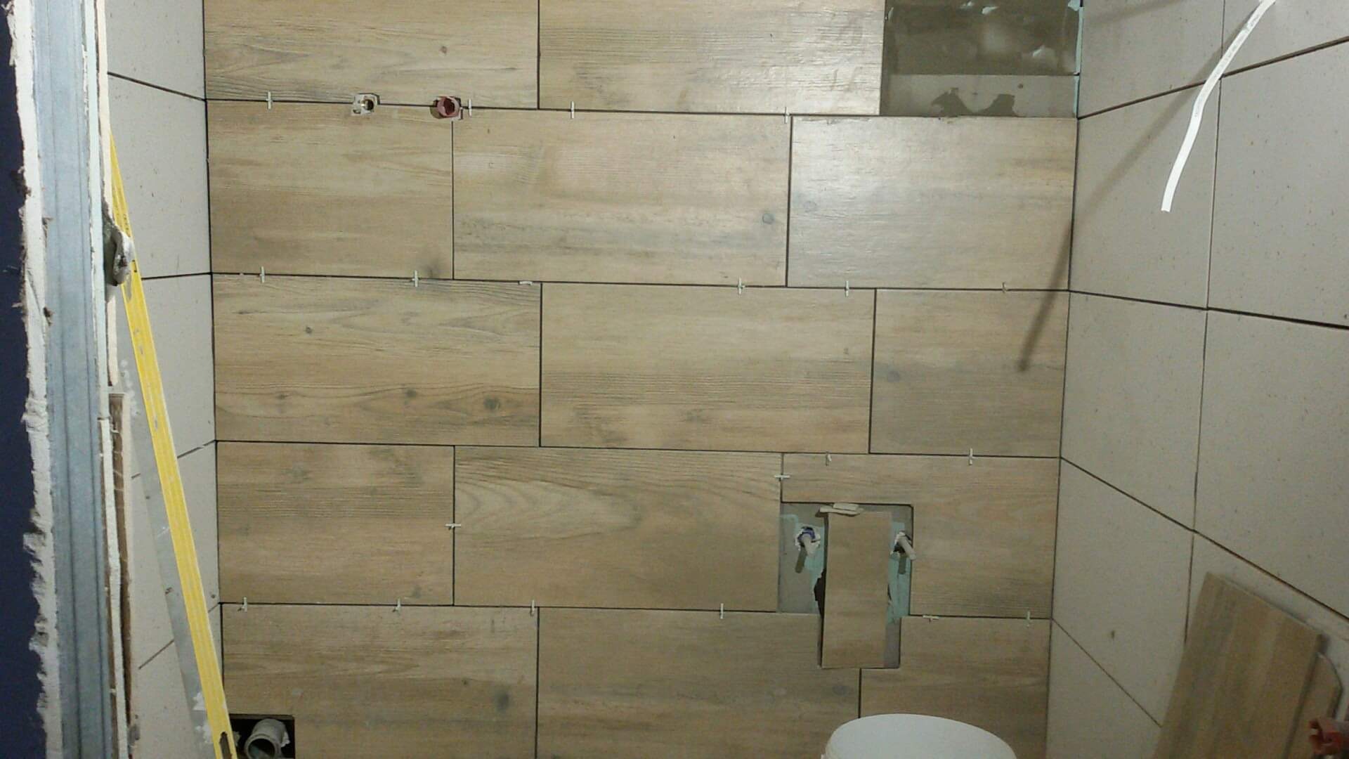 Ściana łazienki w trakcie układania płytek imitujących drewno, widoczne krzyżyki dystansowe, otwory na instalacje i poziomica przy ścianie.