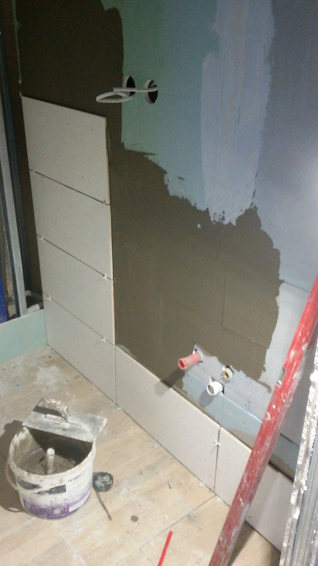 Ściana w trakcie układania płytek ceramicznych, widoczne krzyżyki dystansowe, wiadro z zaprawą klejową oraz fragment instalacji wodnej.
