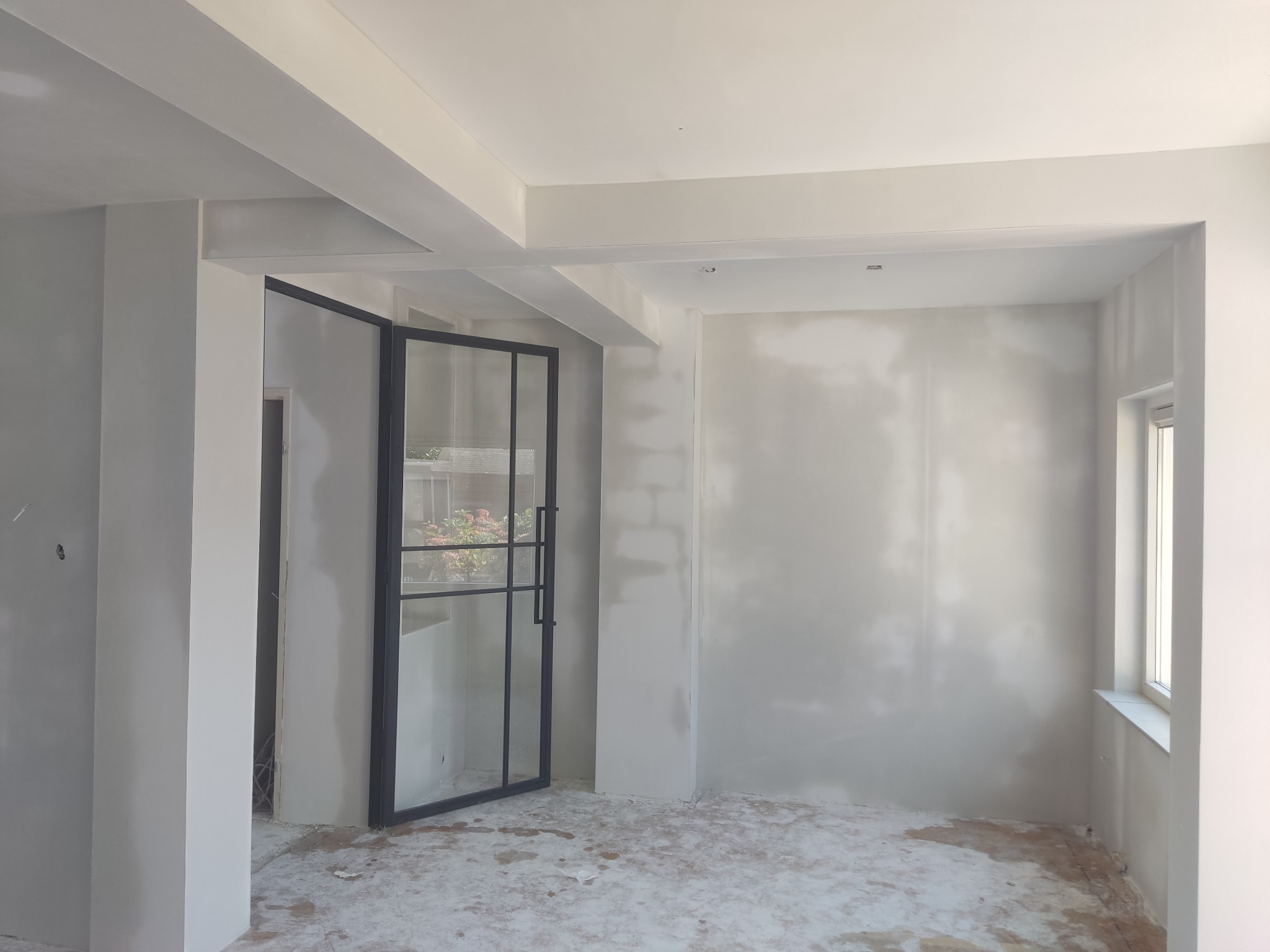 Wnętrze w trakcie prac wykończeniowych: szpachlowane ściany, otwarte czarne drzwi szklane z widokiem na rośliny, surowa podłoga z resztkami gipsu. Widoczne filary i surowe wykończenie sufitu.