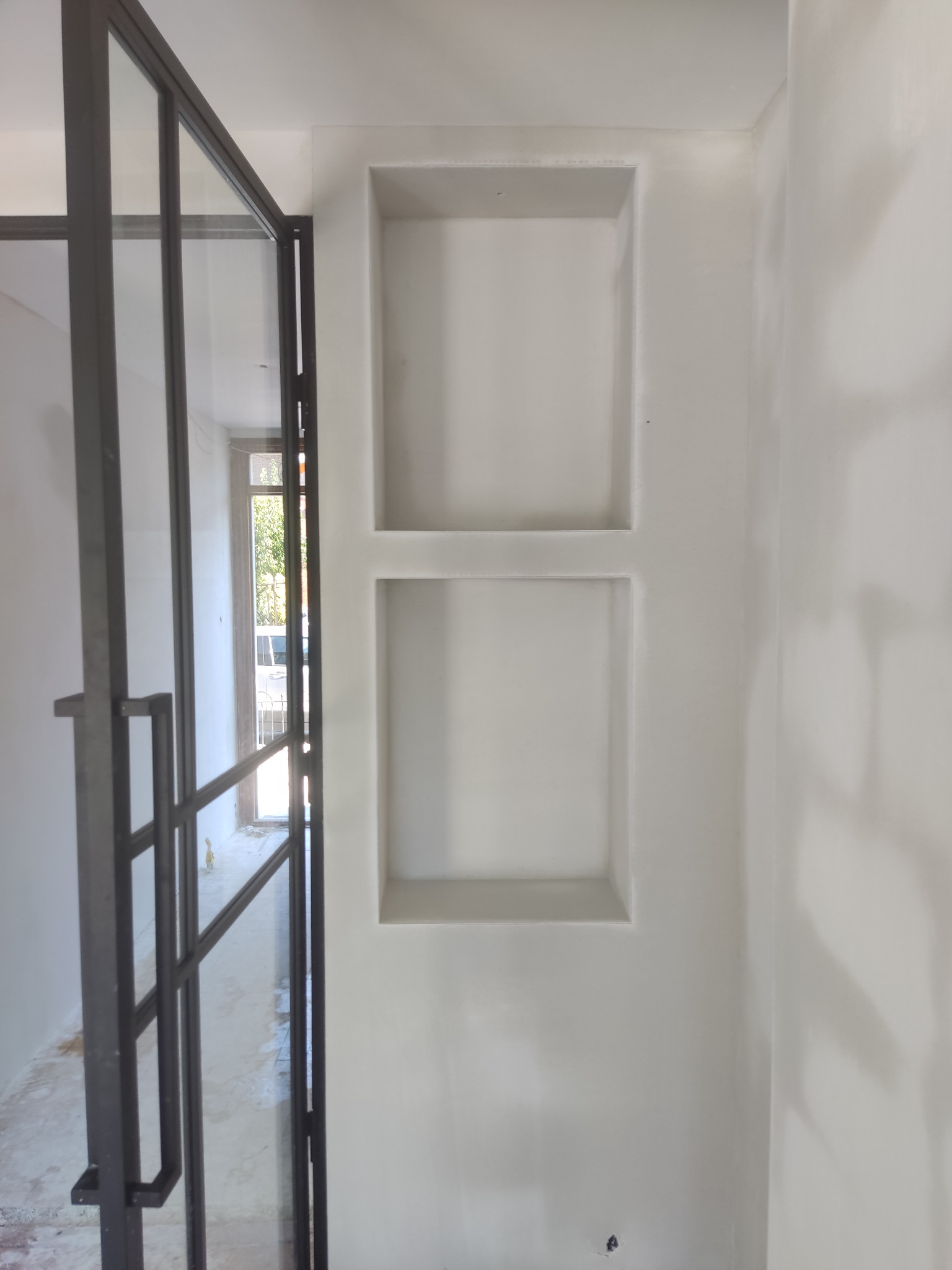 Wnęki ścienne w surowym stanie, z widocznymi śladami po gładzi, obok przeszklonej ścianki działowej w metalowej ramie, podłoga w trakcie prac wykończeniowych.