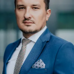 Portret mężczyzny w błękitnym garniturze z wzorzystą poszetką i krawatem w kratę na tle szklanej fasady budynku.