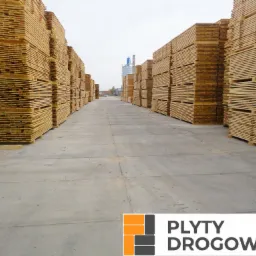 Składowane płyty drogowe z drewna na betonowym placu, widok perspektywiczny w kierunku silosów w tle.