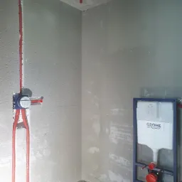 Surowy stan łazienki w trakcie instalacji: widoczne czerwone rury w ścianie, podtynkowa bateria Grohe i stelaż podtynkowy WC Grohe z częściowo zamontowaną spłuczką.