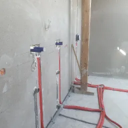 Surowe ściany z widocznymi, czerwonymi rurami instalacji grzewczej w izolacji, wystającymi ze ściany z niebieskimi zaślepkami, szare rury kanalizacyjne na podłodze, drewniany słup w tle, w trakcie...