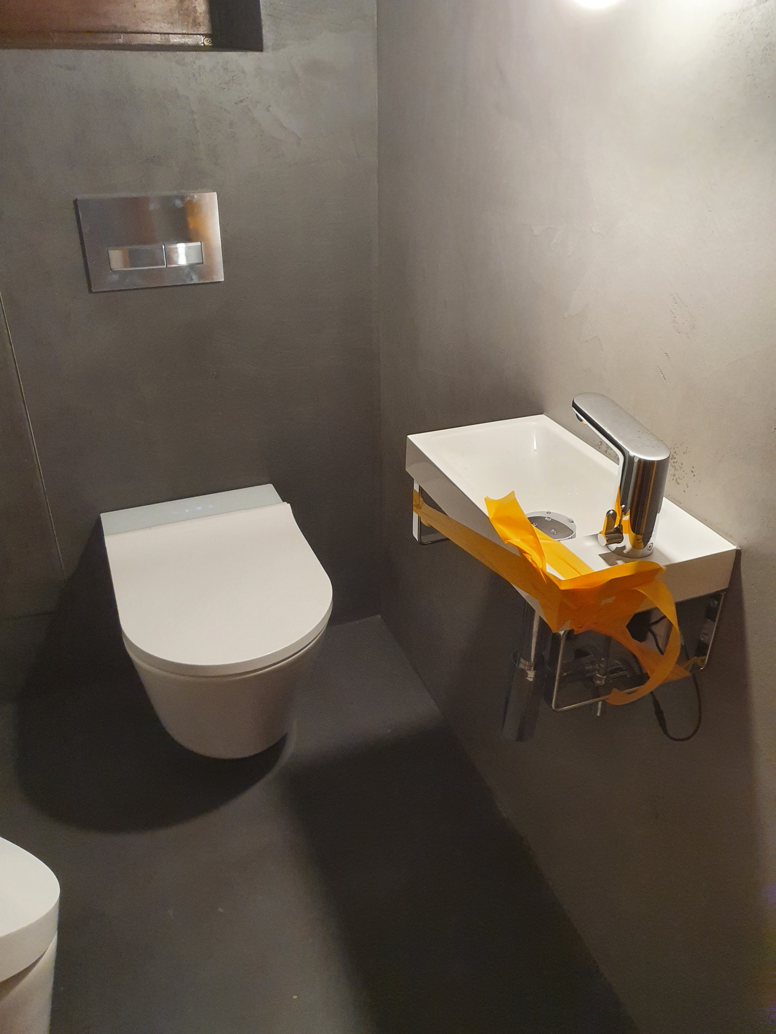 Nowoczesna toaleta wisząca i umywalka z chromowaną baterią, zabezpieczona żółtą taśmą, w łazience wykończonej betonem architektonicznym. Widoczny przycisk spłuczki w ścianie.