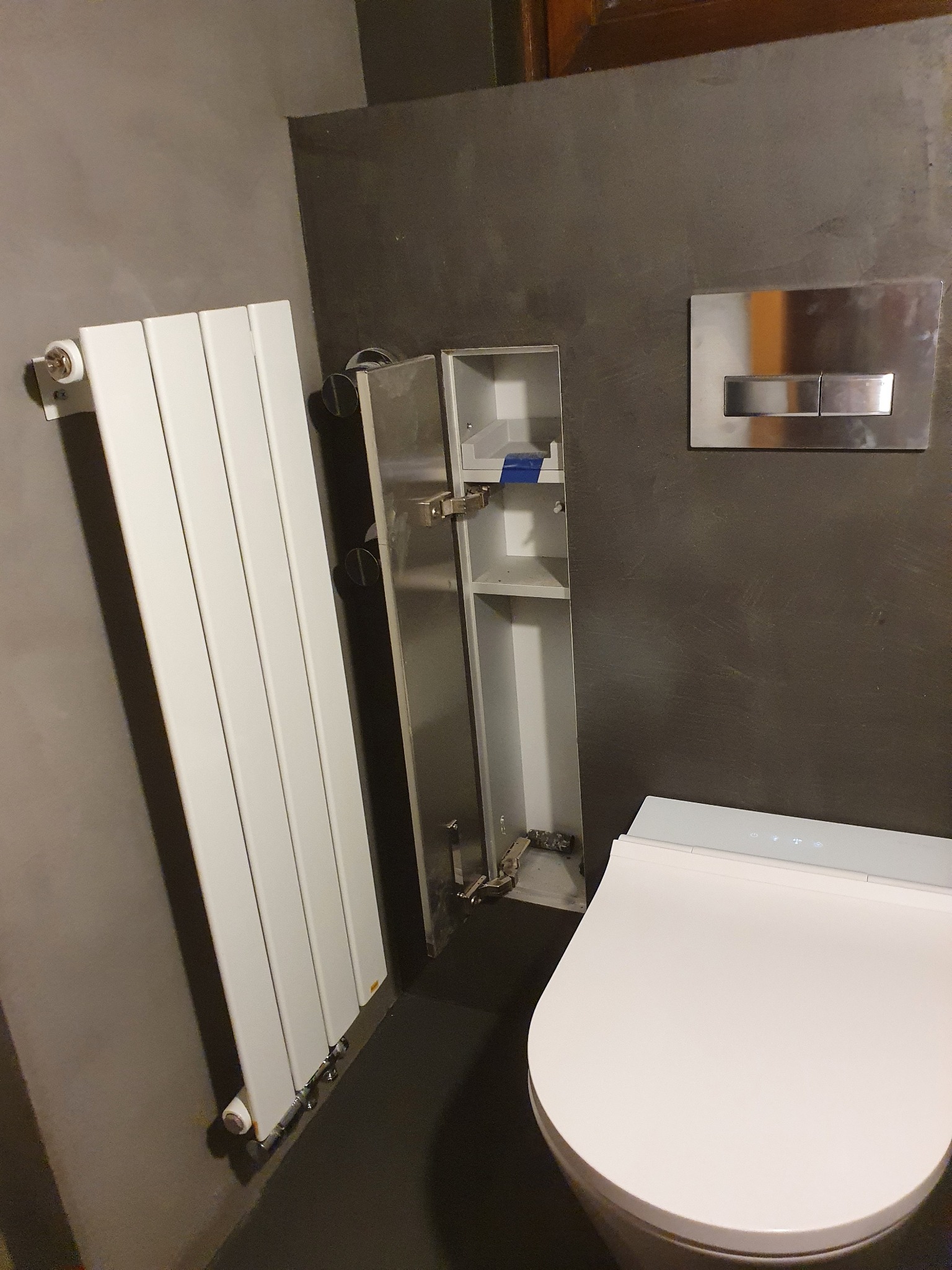 Nowoczesna łazienka z wiszącą toaletą, grzejnikiem panelowym i szafką wnękową z otwartymi drzwiczkami, widoczna instalacja wodno-kanalizacyjna.