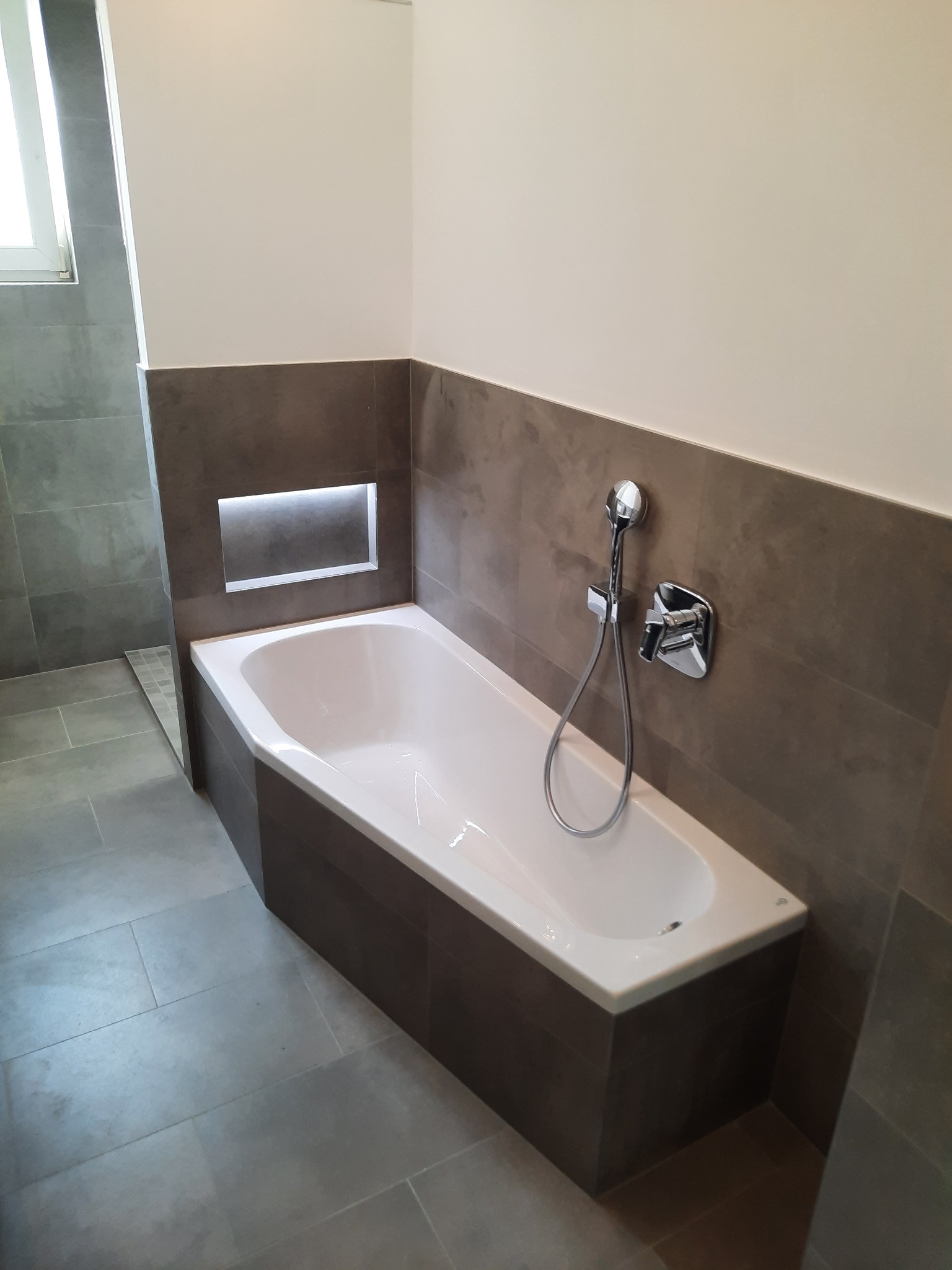 Nowoczesna łazienka z wanną obudowaną ciemnymi płytkami, chromowaną armaturą i wnęką z podświetleniem LED.