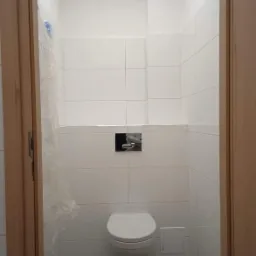 Biała, podwieszana toaleta na tle białych, kwadratowych płytek w łazience, widok przez drewniane futryny drzwi, z lewej strony widoczna folia ochronna.