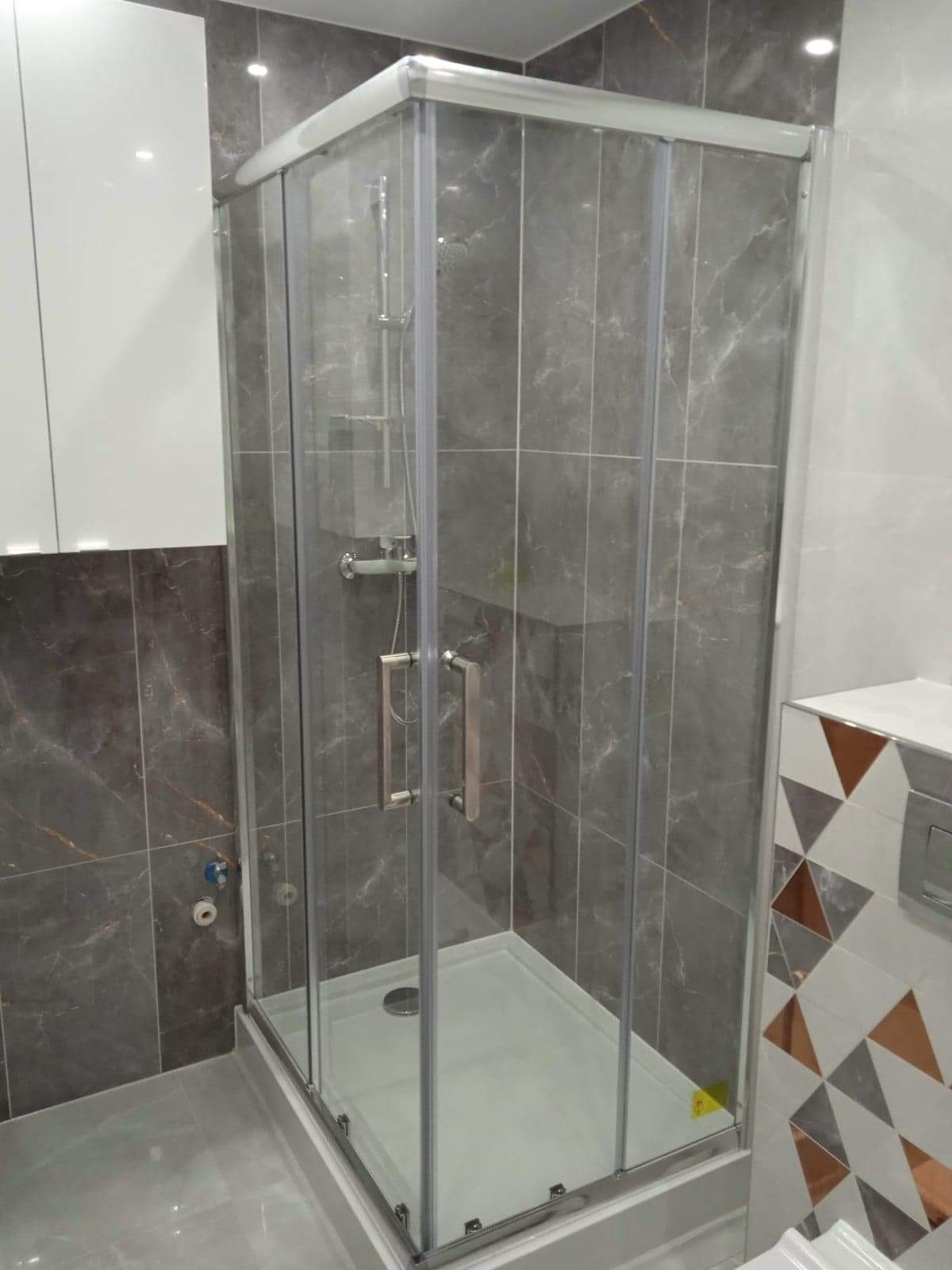 Szklana kabina prysznicowa narożna z przesuwnymi drzwiami, zamontowana w łazience wykończonej szarymi płytkami imitującymi marmur, z widoczną armaturą i odpływem liniowym.