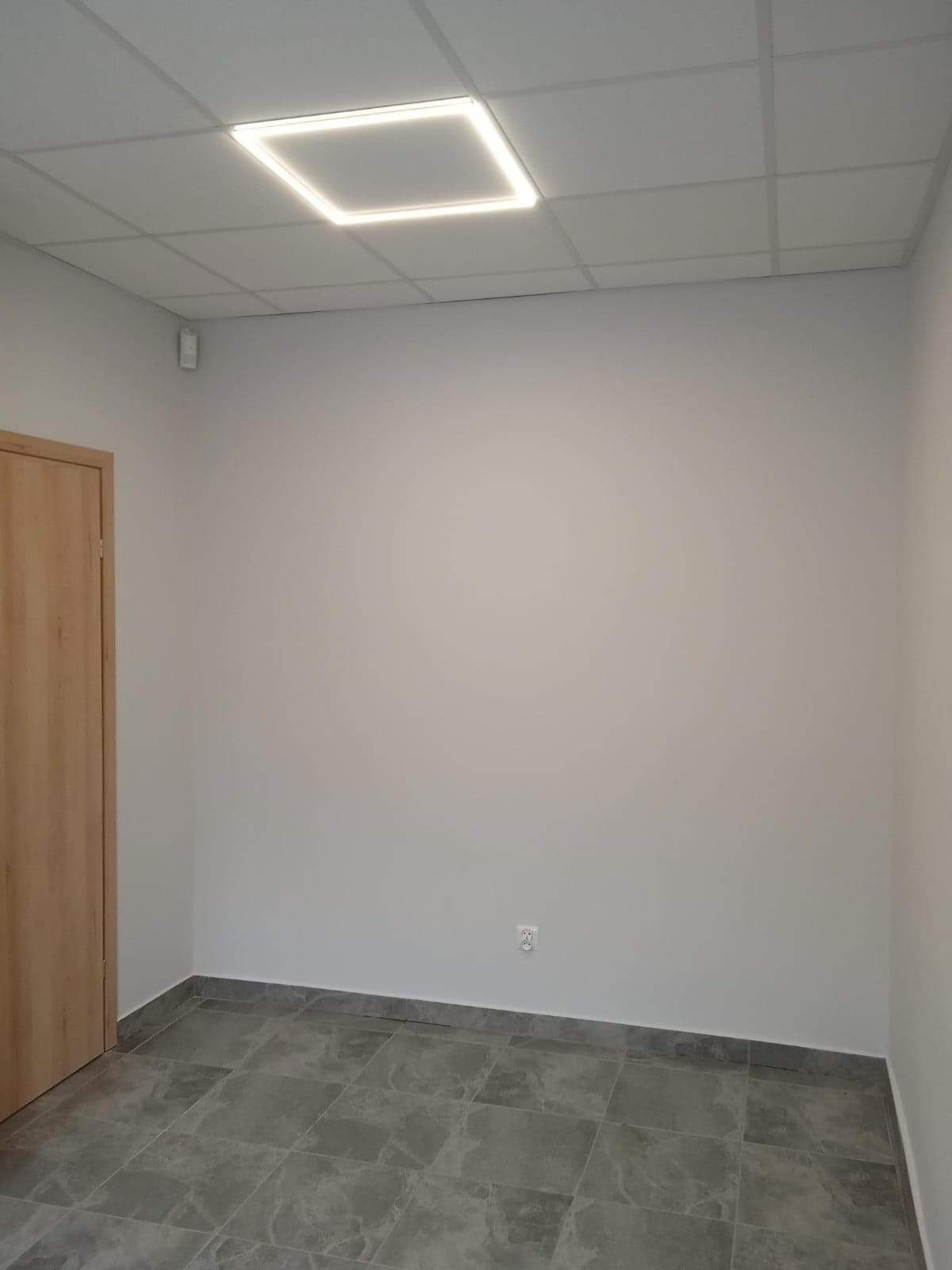 Pusty pokój po remoncie z szarymi płytkami na podłodze, świeżo pomalowanymi ścianami na szaro i nowoczesnym kwadratowym oświetleniem LED w suficie podwieszanym.