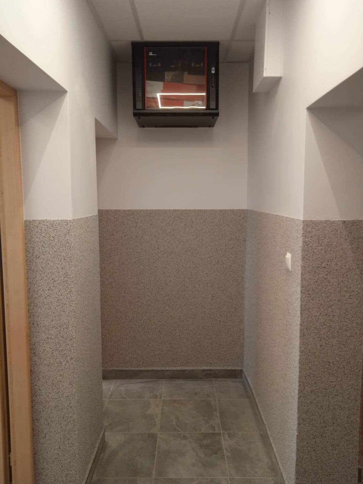 Korytarz z szarą, teksturowaną dolną częścią ścian, białą górną częścią i szarą płytką podłogową, z szafką elektryczną pod sufitem.