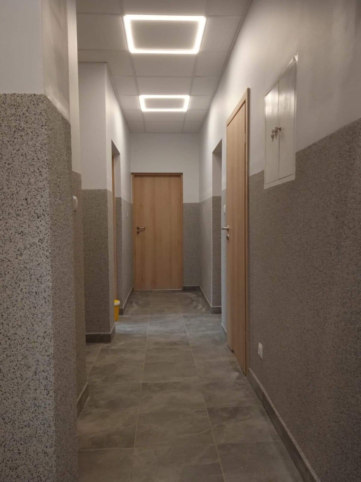 Wnętrze korytarza w budynku mieszkalnym z szarymi płytkami podłogowymi, ścianami pokrytymi tapetą strukturalną w dwóch odcieniach szarości, drewnianymi drzwiami i nowoczesnym oświetleniem sufitowym...