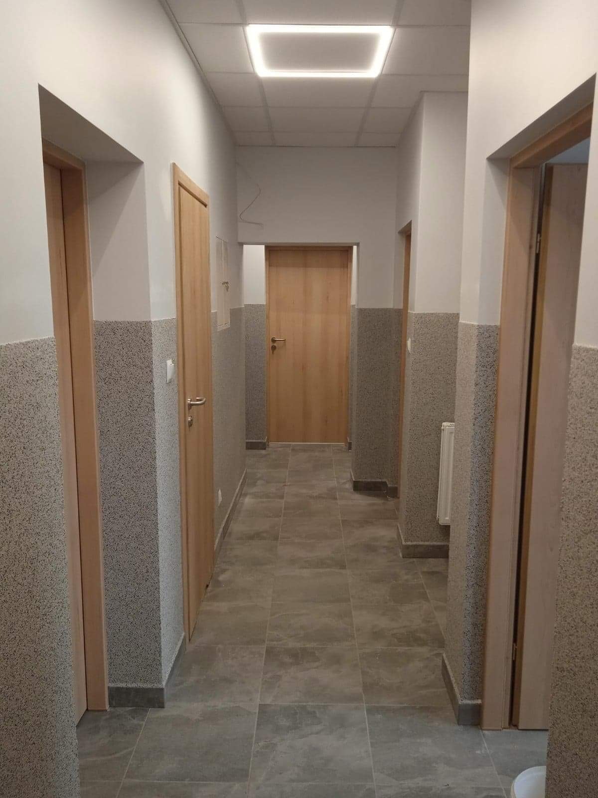Korytarz w budynku z szarymi płytkami na podłodze i ścianach do połowy wysokości, drewniane drzwi, kwadratowa lampa sufitowa.