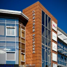 Zewnętrzny widok nowoczesnego budynku biurowego z napisem 'Centrum Biurowe' na elewacji, widoczne okna odbijające niebo i tabliczki firmowe.