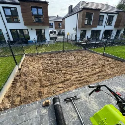 Przygotowany teren pod trawnik na osiedlu domów jednorodzinnych, widoczny walec z siatki, grabie i glebogryzarka, ziemia wyrównana z widocznymi śladami opon.