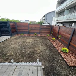 Świeżo założony trawnik z ciemną ziemią, obrzeża z kory i kamienia, drewniany płot z metalowymi słupkami, nowoczesna architektura budynku w tle.