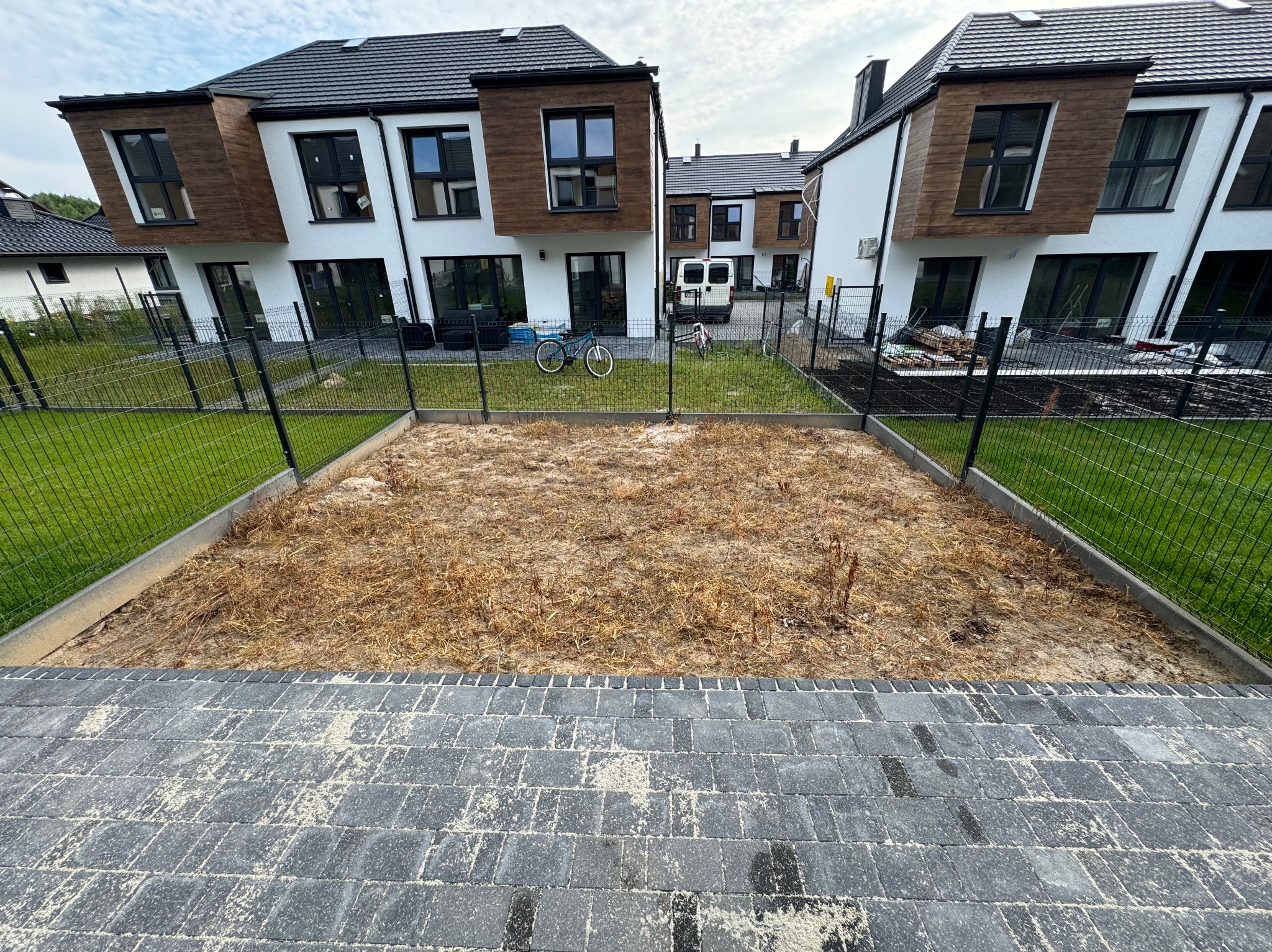 Przygotowany teren pod trawnik z widocznymi pozostałościami starej trawy, ogrodzony siatką, w tle nowoczesne domy i rowery.