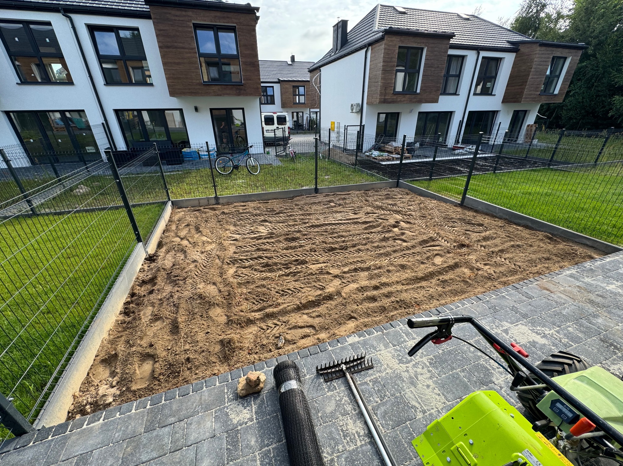 Przygotowany teren pod trawnik na osiedlu domów jednorodzinnych, widoczny walec z siatki, grabie i glebogryzarka, ziemia wyrównana z widocznymi śladami opon.