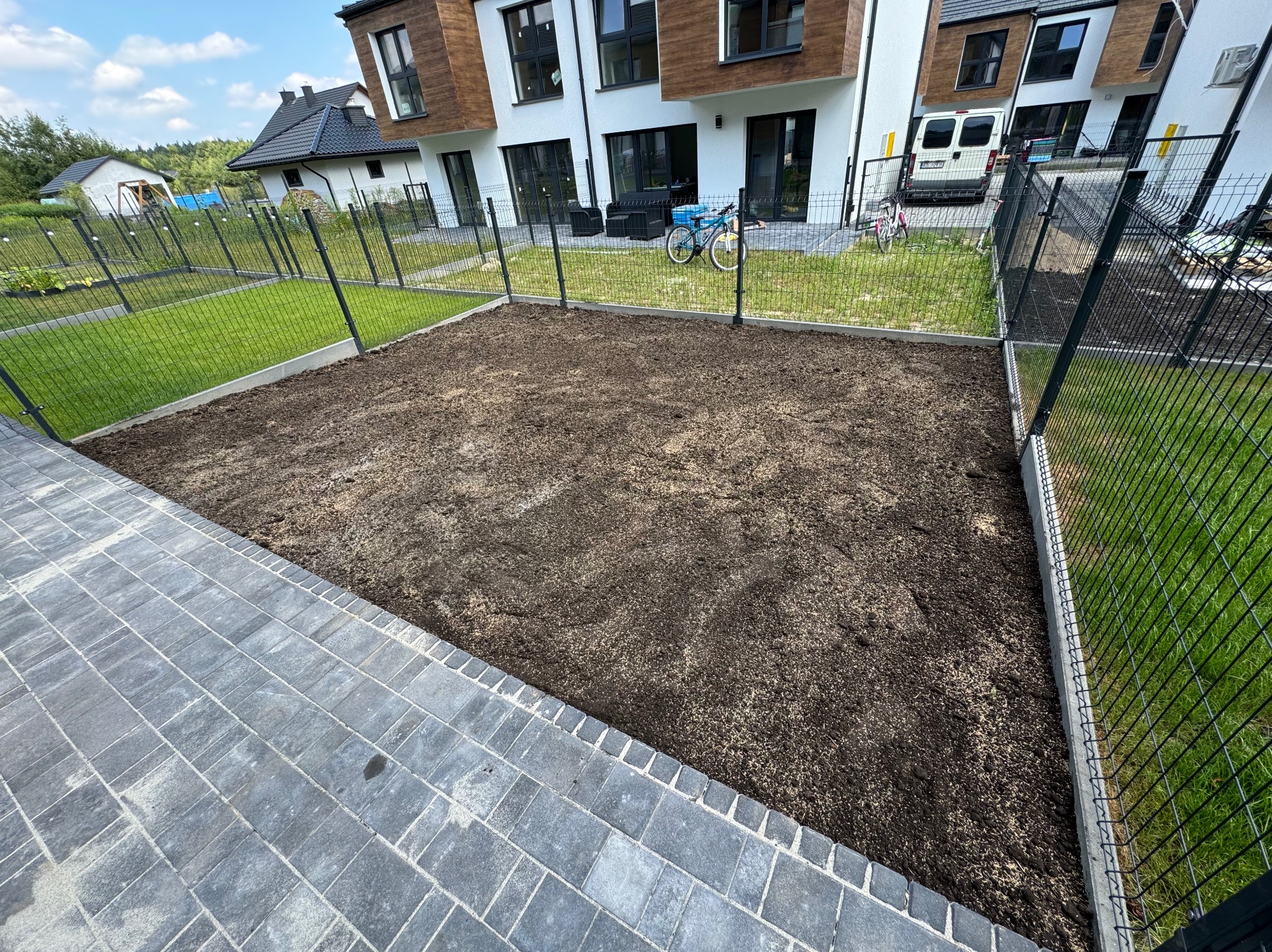 Przygotowany teren pod trawnik z widoczną warstwą ziemi, obrzeżami i ogrodzeniem panelowym na tle nowoczesnych domów.