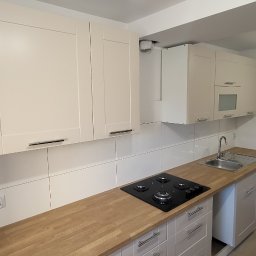 NikalayeuRO - Nowoczesna kuchnia z jasnymi, kremowymi szafkami, drewnianym blatem, płytą grzewczą i metalowym zlewem, widok na fragment pomieszczenia z mopem opartym o ścianę.