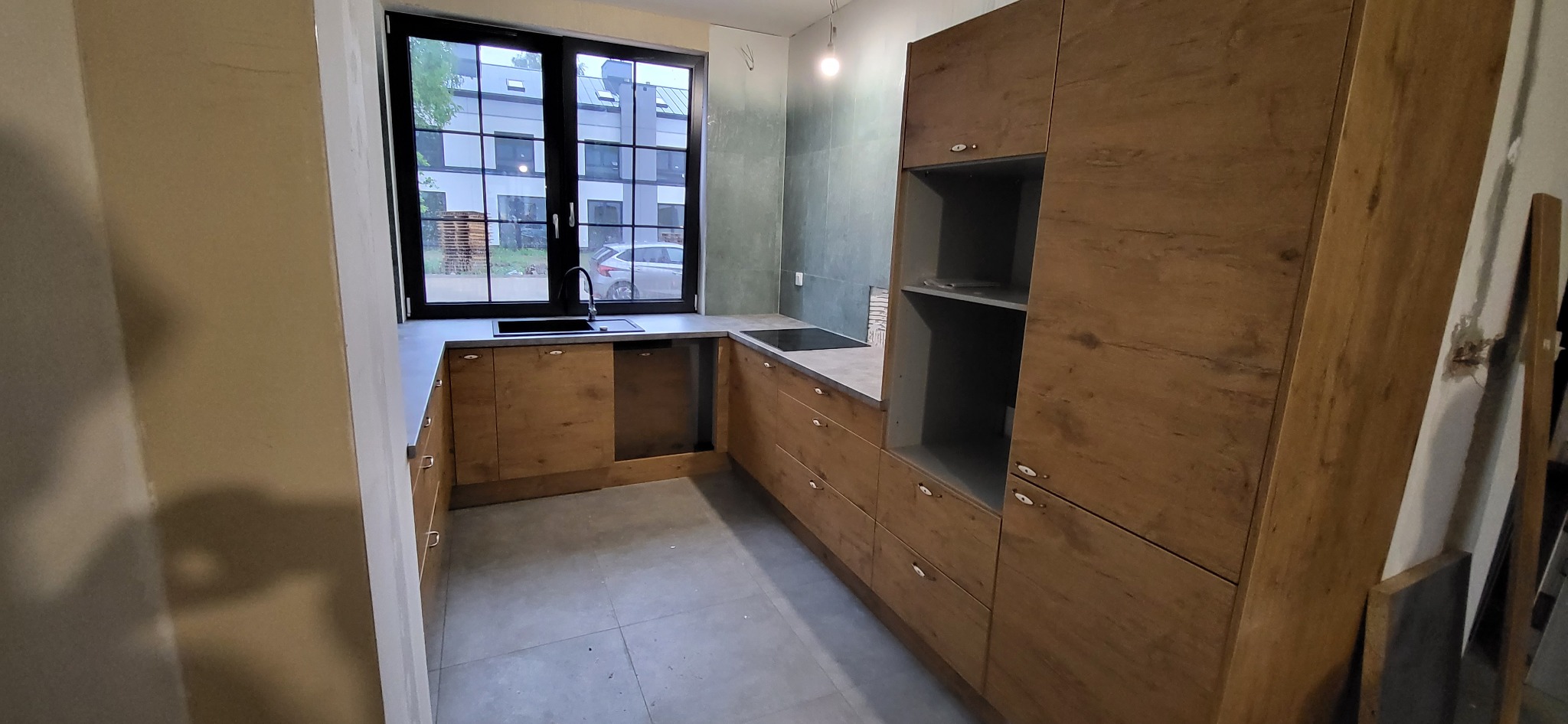 Kątowe ustawienie drewnopodobnych szafek kuchennych z szarym blatem i czarnym zlewem pod oknem w surowym wnętrzu.