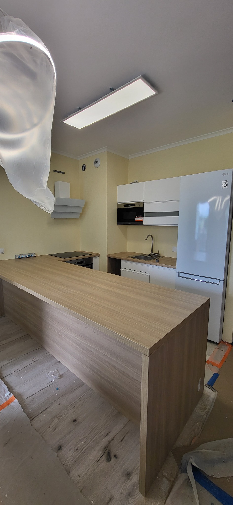 Montaż kuchni: wyspa kuchenna z blatem drewnianym w trakcie instalacji, widoczne zabezpieczenia podłogi i folia ochronna na elementach kuchni.