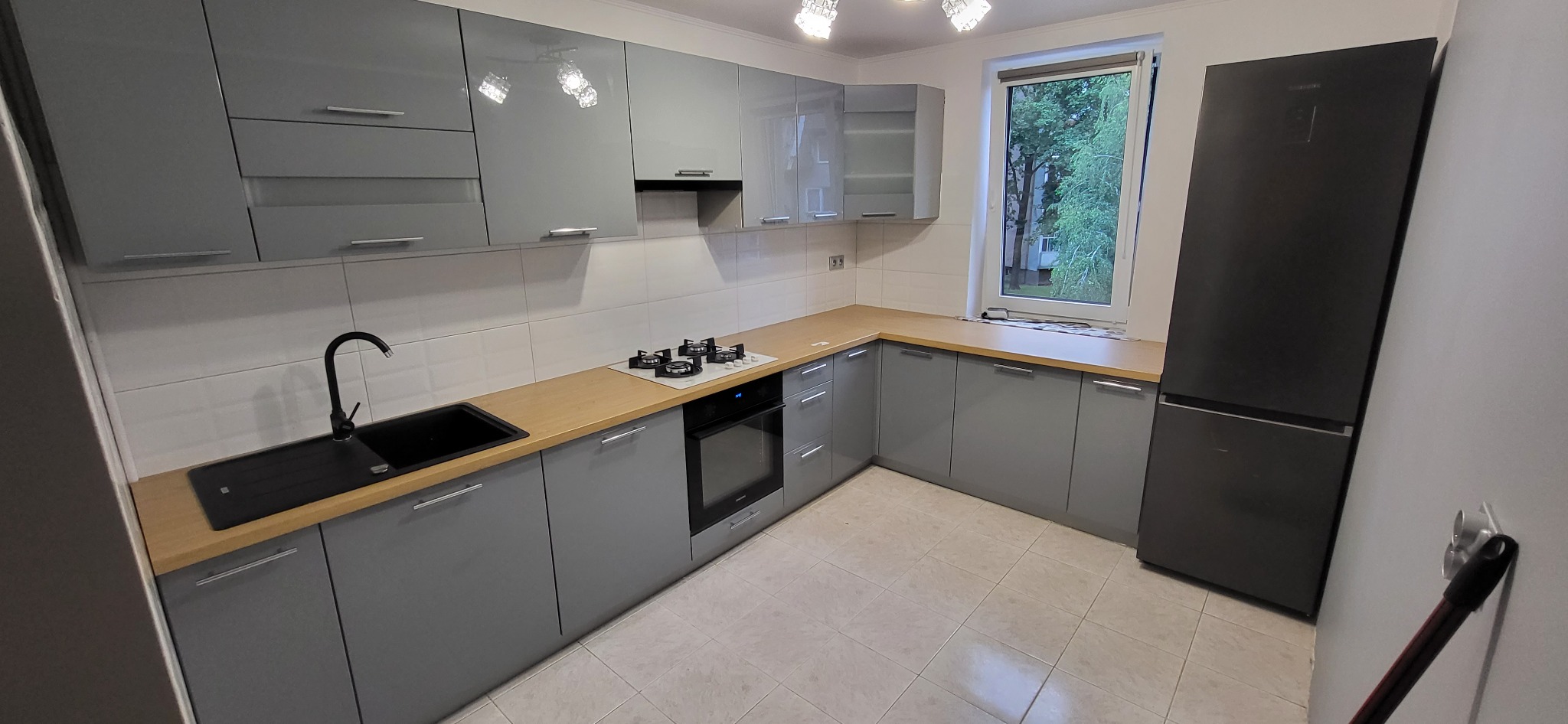 Szara kuchnia z drewnianym blatem, czarnym zlewem i czarną lodówką, oświetlona lampą sufitową z wieloma żarówkami.