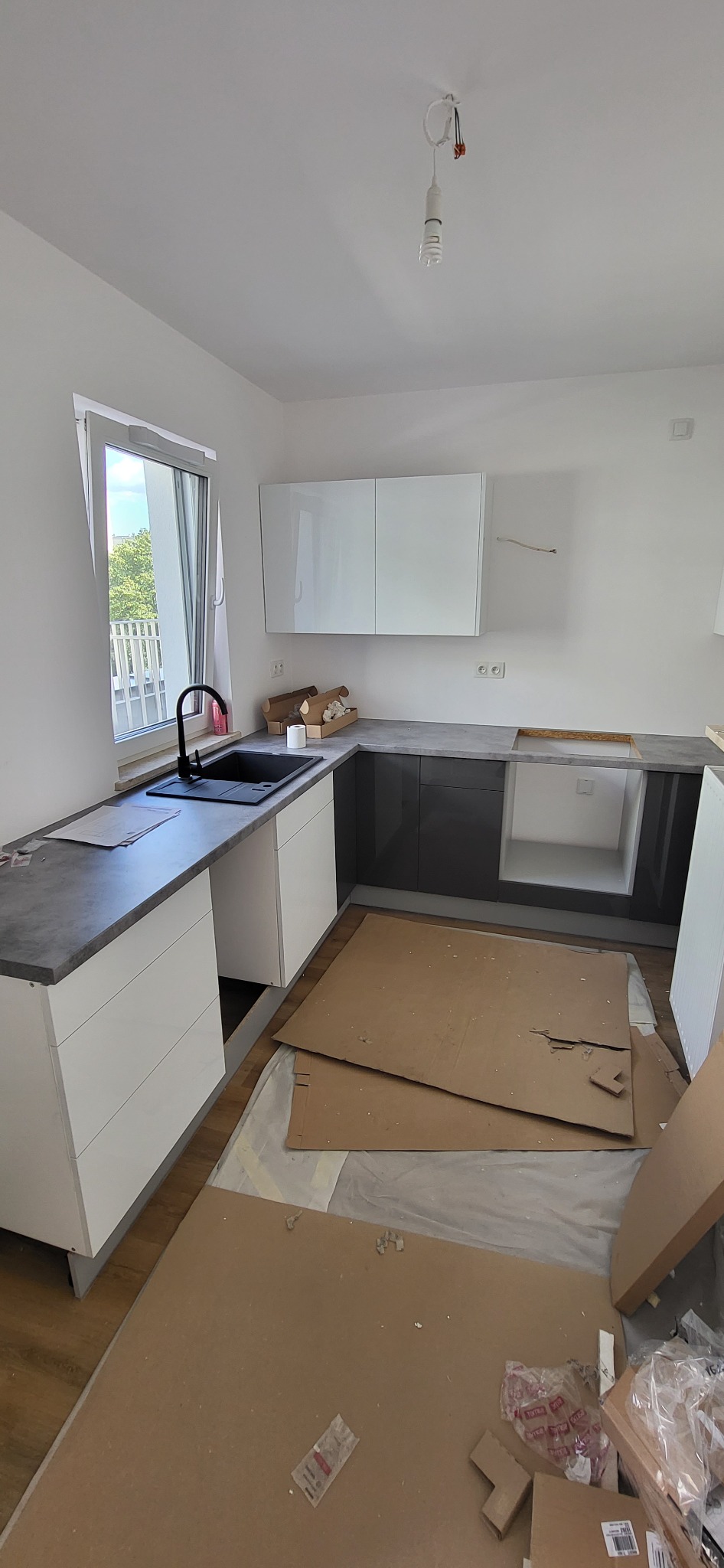 Montaż szarego blatu na białych i grafitowych szafkach kuchennych, widoczne kartony zabezpieczające podłogę, surowa żarówka na kablu.