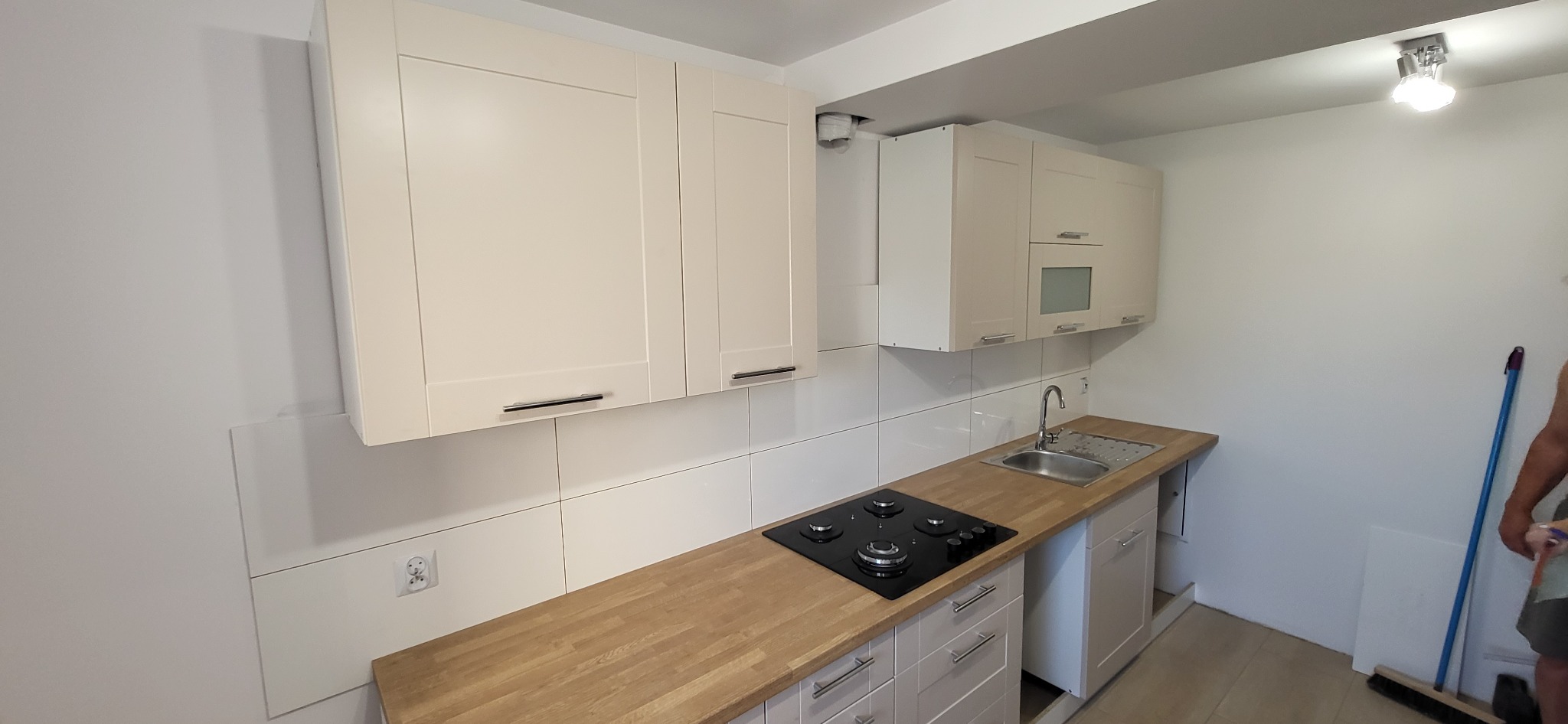 Nowoczesna kuchnia z jasnymi, kremowymi szafkami, drewnianym blatem, płytą grzewczą i metalowym zlewem, widok na fragment pomieszczenia z mopem opartym o ścianę.