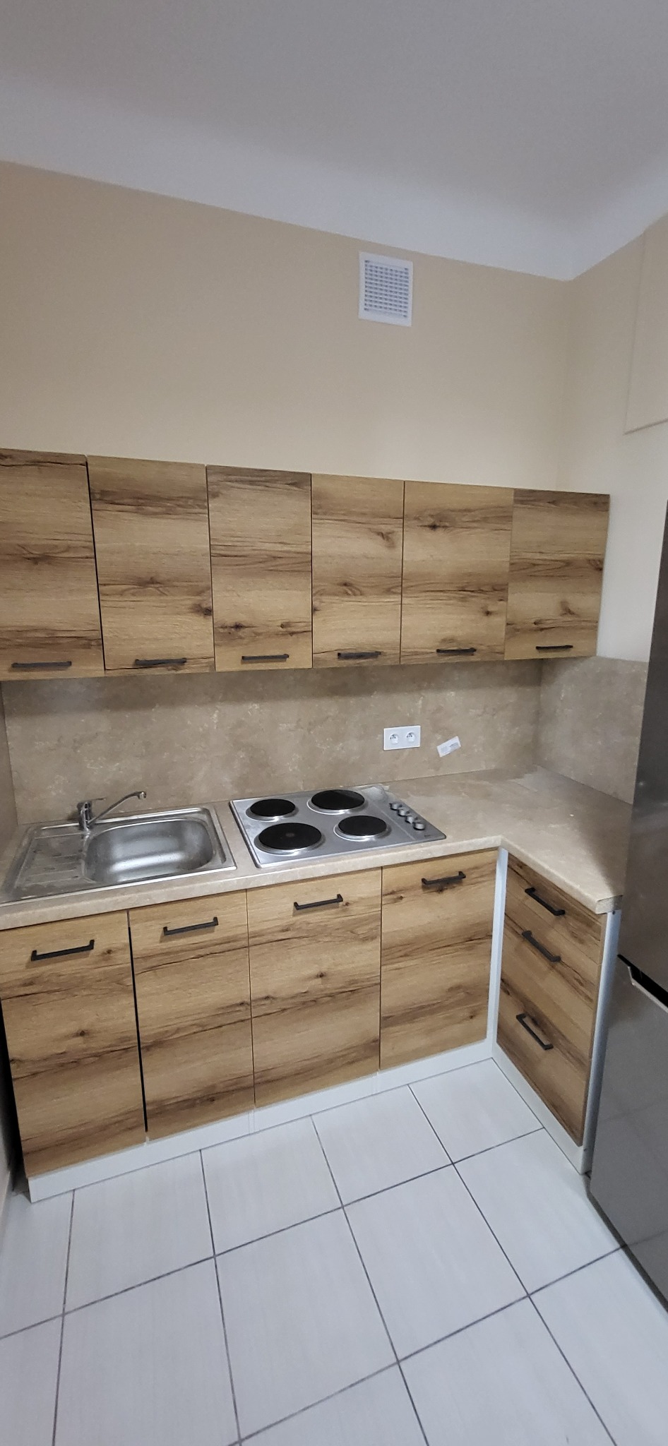 Kuchenna zabudowa z drewnopodobnymi frontami szafek, czarne uchwyty, jasny blat i płytki podłogowe, widok z perspektywy narożnika pomieszczenia.