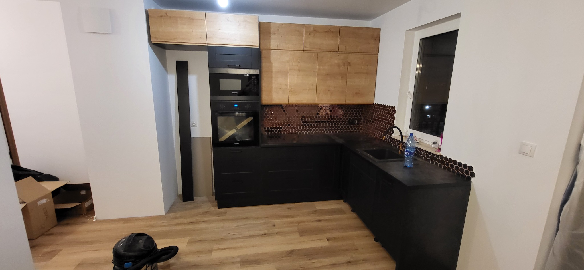 Częściowo ukończona kuchnia: czarne szafki dolne, drewniane górne, mozaika heksagonalna na ścianie, widoczny sprzęt AGD i kartony na podłodze.